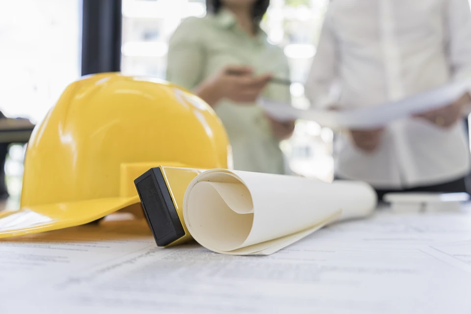 Scène de bureau d'architecture ou d'ingénierie avec casque de chantier jaune, plans roulés et tampon encreur sur table blanche