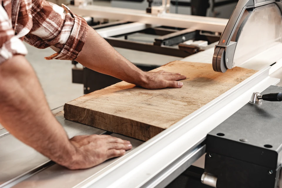 Photographie d'un atelier de menuiserie professionnel avec deux ou trois menuisiers travaillant sur un agencement de cuisine en bois massif. Au premier plan, un artisan mesure ou assemble une partie de meuble. Un collègue travaille en arrière-plan. L'atelier doit montrer l'équipement et le savoir-faire : bois de qualité, outils de précision, ambiance de travail structurée et organisée. La lumière naturelle doit valoriser le bois et créer une atmosphère professionnelle et accueillante.