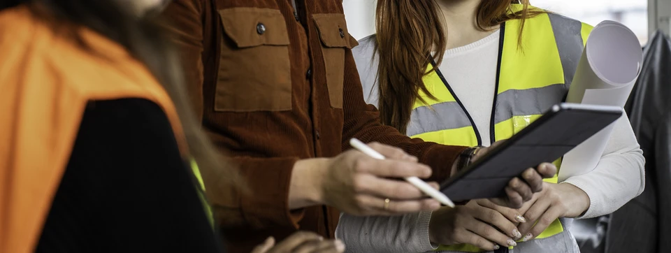 Photographie d'un professionnel en tenue de chantier (gilet de sécurité) sur un chantier de construction immobilier moderne, tenant des plans ou une tablette, en discussion avec un autre ouvrier ou responsable de projet. Le contexte montre clairement un environnement de bâtiment professionnel en cours de réalisation, avec structures et éléments en bois visibles. L'image doit communiquer la coordination, la responsabilité et le suivi méthodique d'un projet collectif.
